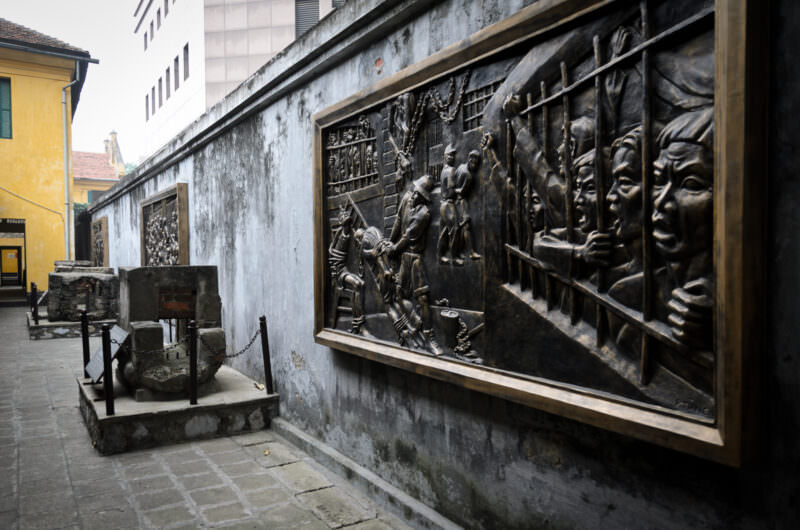 Bronze relief murals beside a preserved sewer tunnel in a Hoa Lo Prison courtyard