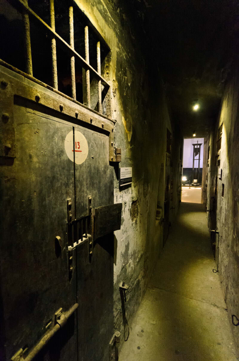 Iron door numbered 13 sealing a death row cell along a corridor toward a guillotine at Hoa Lo Prison