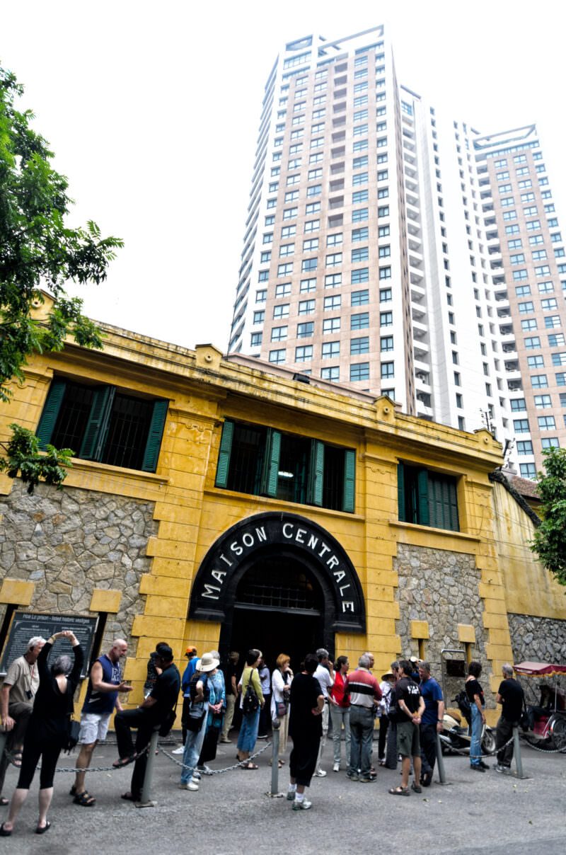 Tourists outside the Maison Centrale gatehouse of Hoa Lo Prison beneath a modern high-rise