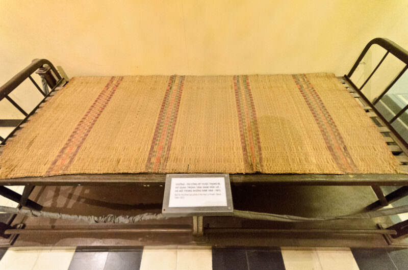 Metal bed with a woven mat displayed as a POW-era artifact at Hoa Lo Prison