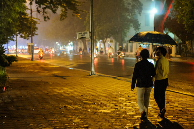 Two women sharing an umbrella on a rain-slicked sidewalk beside Hoan Kiem Lake at night