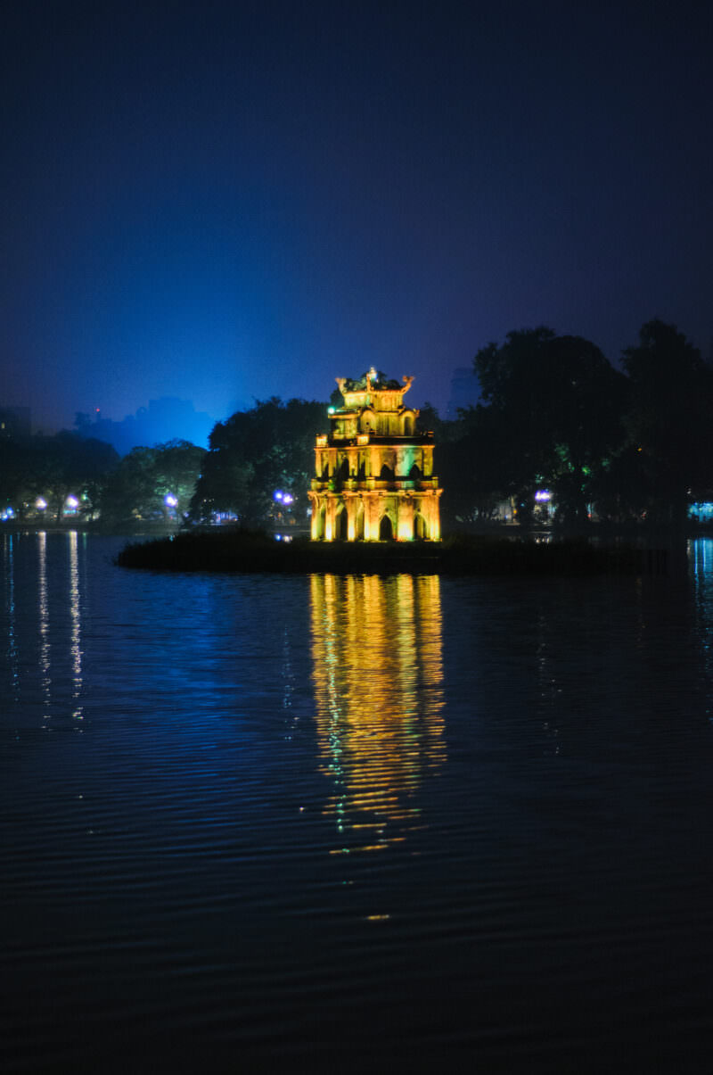 Illuminated Turtle Tower glowing gold on its islet against a deep blue night sky over Hoan Kiem Lake