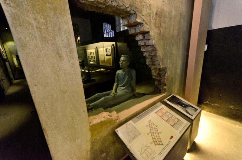 Diorama of a shackled inmate seen through a cutaway brick wall in a Hoa Lo Prison death row cell