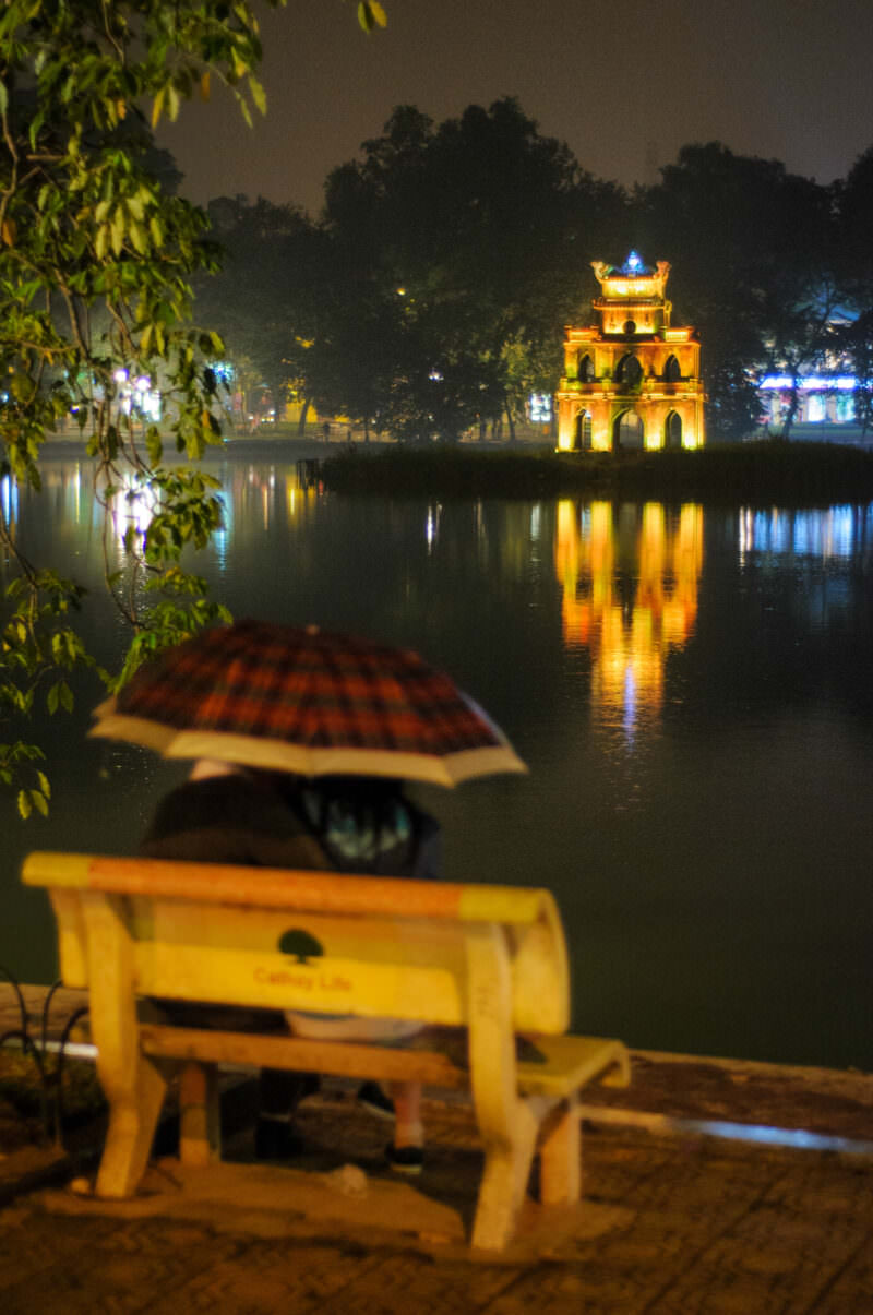 Person under an umbrella on a bench facing the floodlit Turtle Tower across Hoan Kiem Lake