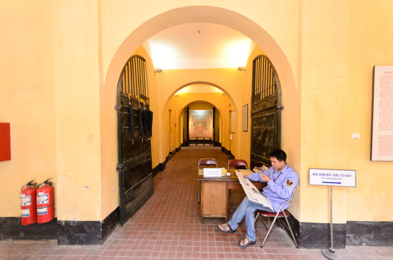 Guard reading a newspaper at the arched iron-gated entrance of Hoa Lo Prison Museum