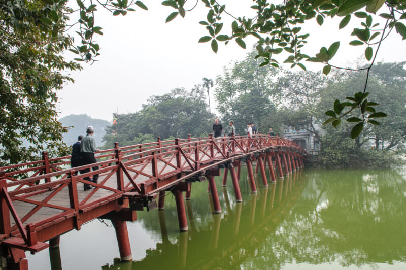 Pedestrians crossing the red Thê Húc Bridge toward Ngoc Son Temple on a misty day