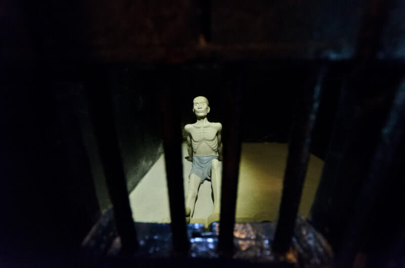 Emaciated prisoner sculpture viewed through iron bars in a Hoa Lo Prison solitary cell
