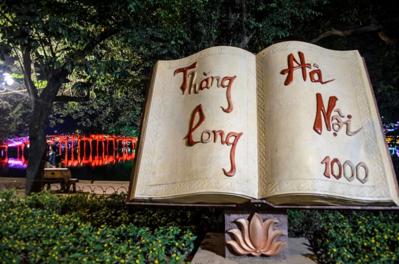Illuminated open-book Thang Long sculpture on the Hoan Kiem Lake shore with The Huc Bridge glowing behind
