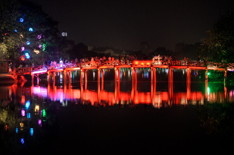 Crowds crossing the red-lit Huc Bridge, its long reflection stretching across Hoan Kiem Lake