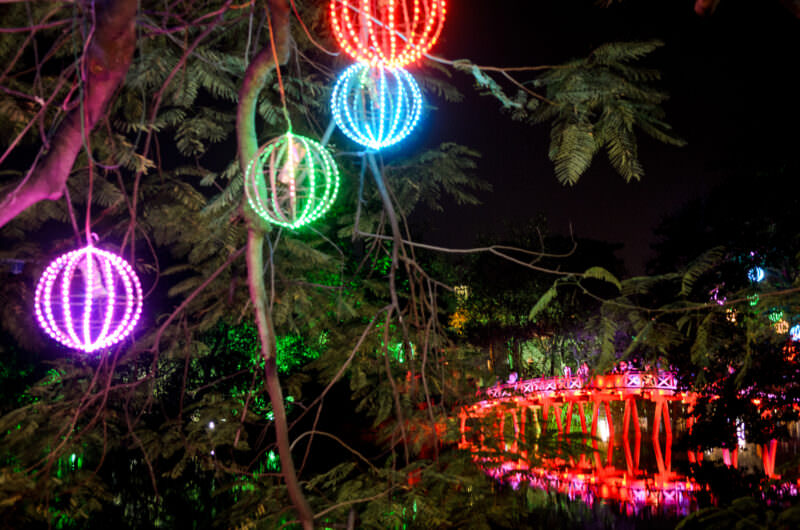 Colorful lantern globes hanging from lakeside trees with the red Huc Bridge glowing beyond