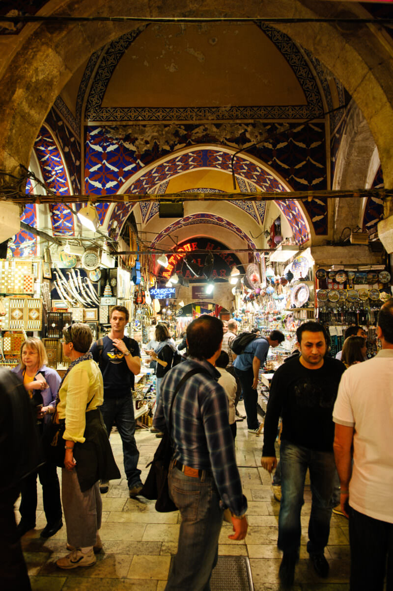 Shoppers walking through a Grand Bazaar corridor beneath an ornately painted geometric vaulted ceiling