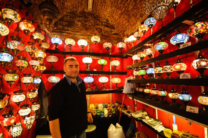 Merchant standing beneath a stone-vaulted ceiling amid shelves of mosaic glass lamps in the Grand Bazaar