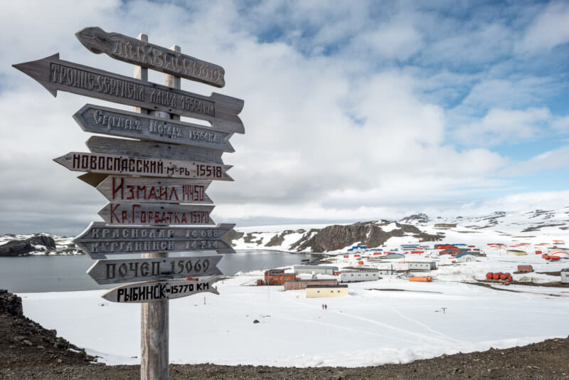 Wooden signpost displaying distances to Russian cities with Bellingshausen and Frei bases spread across Fildes Peninsula below, King George Island