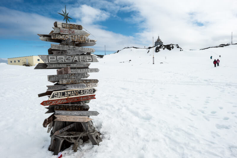 Wooden signposts showing distances to cities with Trinity Church visible on rocky hill behind at Bellingshausen Station, King George Island