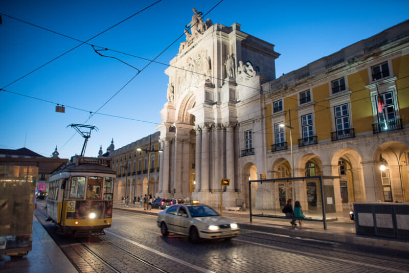 Arco da Rua Augusta illuminated at dusk with a yellow tram and taxi passing on Praça do Comércio