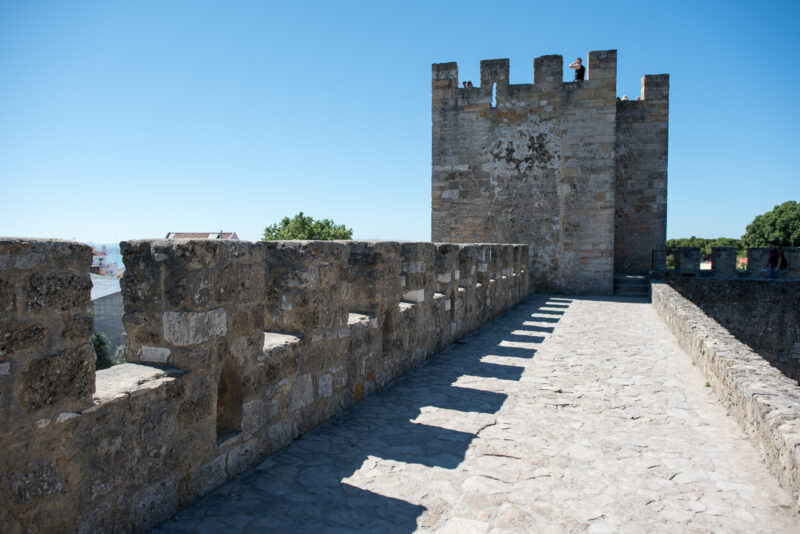 A square stone tower rises above the crenellated ramparts of Castelo de São Jorge in bright sunlight
