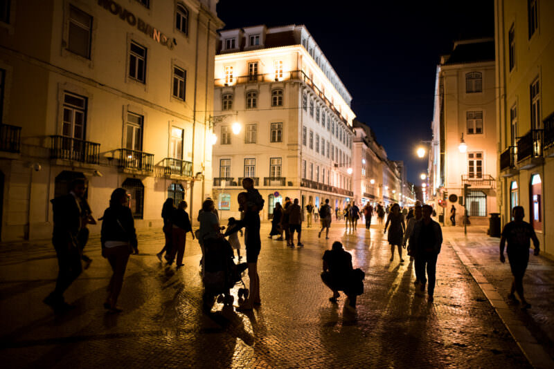Silhouetted pedestrians and families crossing Rua Augusta at night beneath glowing Pombaline facades