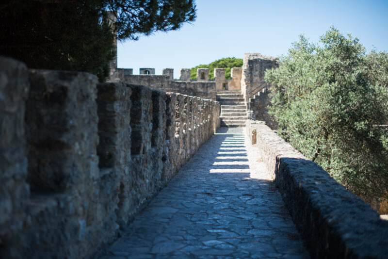 A shadow-striped stone walkway runs along the ramparts of Castelo de São Jorge toward a stairway
