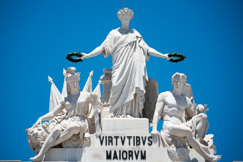 Close-up of the allegorical marble figures crowning the Arco da Rua Augusta above the Latin inscription