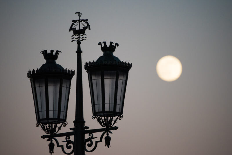 Ornate ship-crested street lamp silhouetted against a full moon on Praça do Comércio in Lisbon