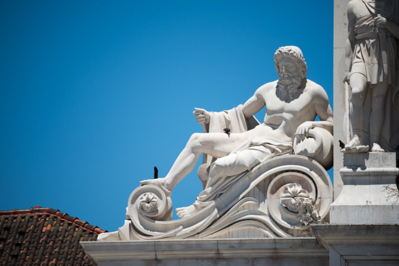 Reclining allegorical stone figure atop the Arco da Rua Augusta set against a clear blue sky