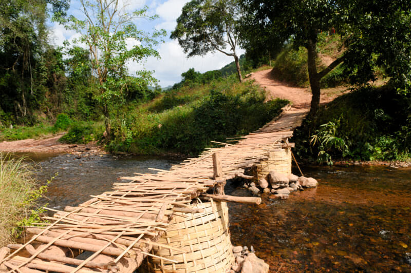 Woven bamboo bridge with stone-filled basket piers crossing a shallow creek, dirt paths and forested hills beyond