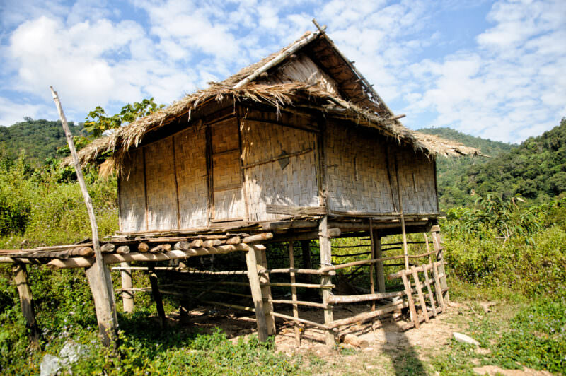 Woven bamboo rice storage hut on tall stilts with thatched roof, forested mountains under cloudy sky behind