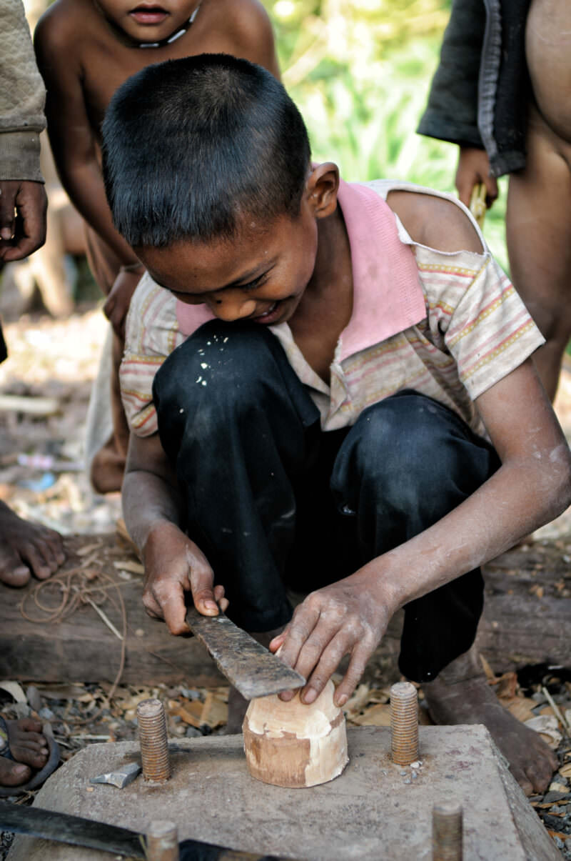 Boy bent over a concrete slab, using a blade to shape a wooden block with metal bolts visible