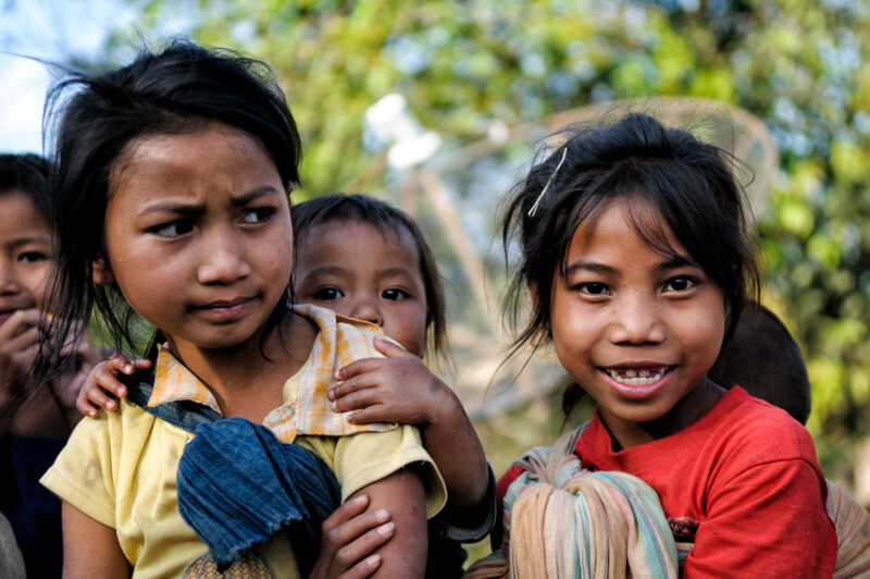 Young girls carrying infants in cloth slings, standing before a mesh satellite dish with children around them