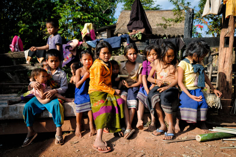 Girls in colorful traditional sinh skirts sitting on a low wall, thatched-roof houses and laundry behind