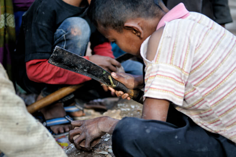 Boy in striped shirt using a machete to carve a wooden block, other children visible around him