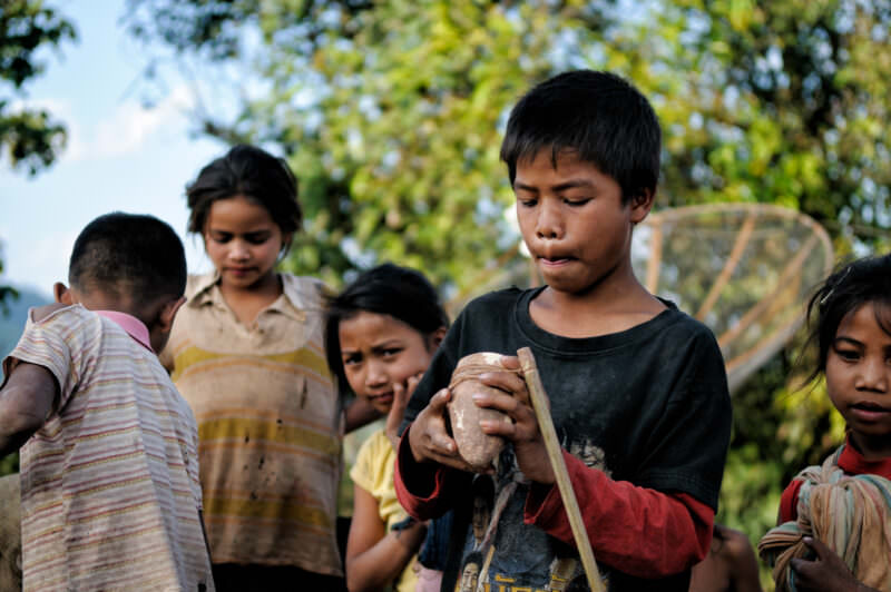 Boy wrapping string around a wooden top, other children and forested mountains visible behind