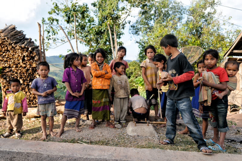 Children gathered on grass with stacked firewood and satellite dish behind, boy holding wooden top and whip
