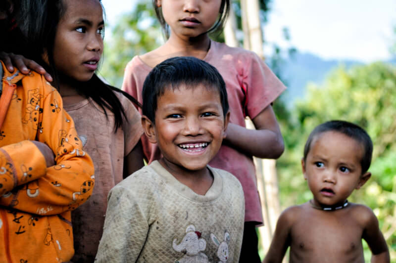 Smiling boy in beige shirt among other children, forested mountain slopes visible behind under blue sky