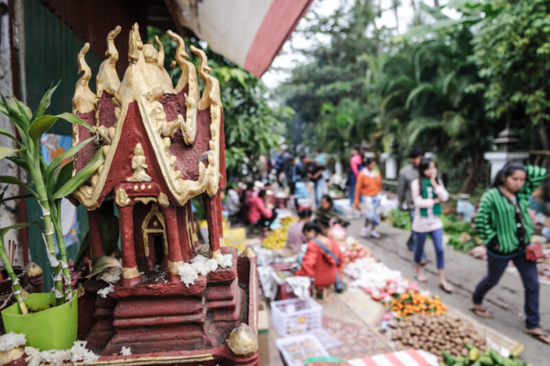 Red and gold spirit house shrine in foreground, its tiered roof sharp against the blurred crowd of Luang Prabang's morning market
