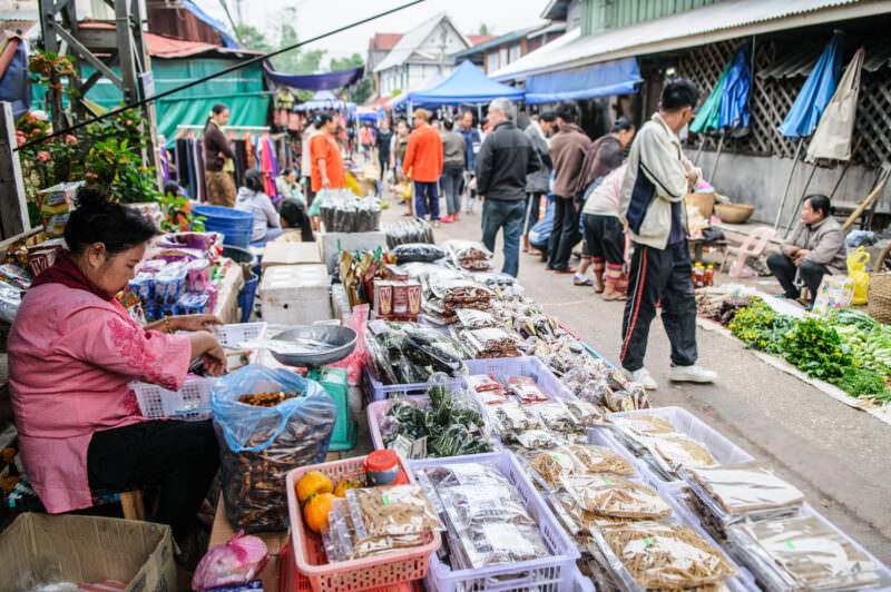 Vendor in pink beside street display of packaged goods and square kaipen sheets, shoppers and blue tarps in background