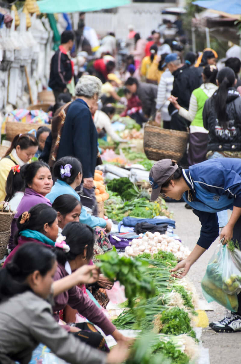 Shoppers crouched over scallions and garlic on ground mats, woven baskets and colorful clothing in a crowded Luang Prabang market lane