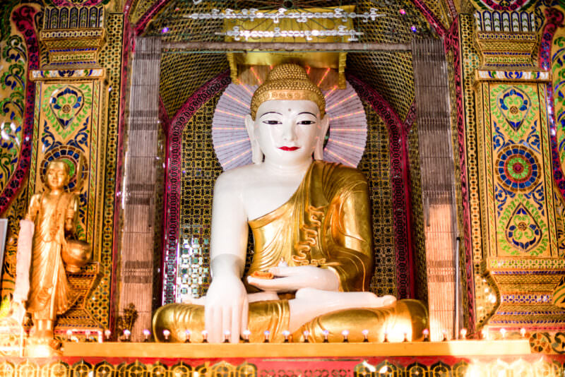 White-and-gold Buddha in bhumisparsha mudra at Sutaungpyei Pagoda, surrounded by mirrored-glass mosaic walls and gold attendant figure
