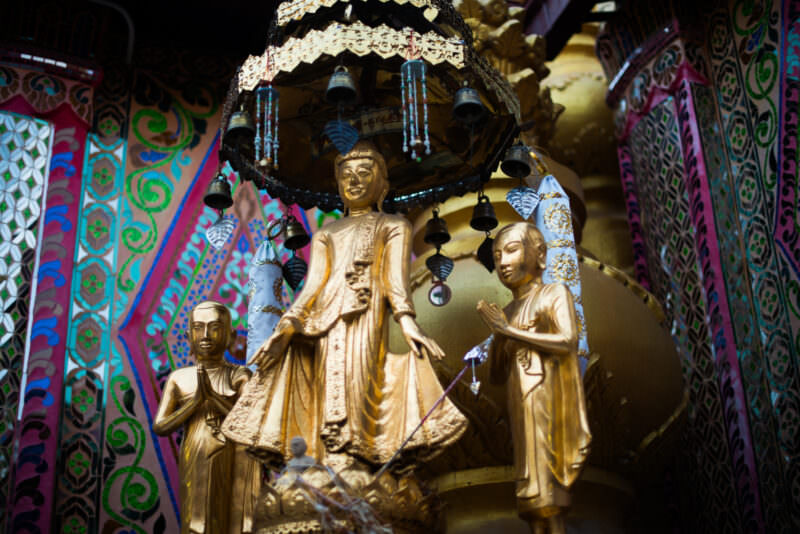 Gilded standing Buddha flanked by two kneeling attendants beneath bell-hung ceremonial umbrella, mirrored-glass mosaic walls at Sutaungpyei Pagoda