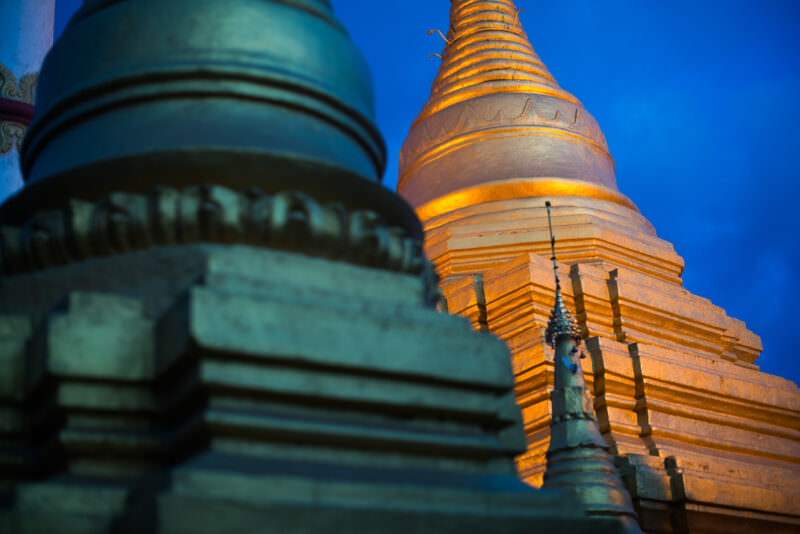 Illuminated main stupa of Sutaungpyei Pagoda glowing against blue-hour sky, smaller stupa in foreground at Mandalay Hill summit