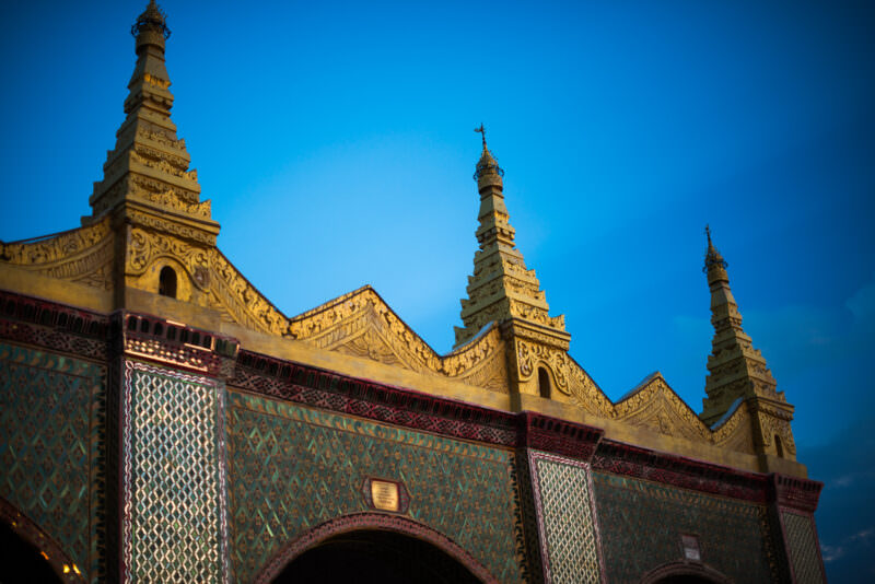 Gilded spires and mirrored-glass mosaic crowning roofline of Sutaungpyei Pagoda at Mandalay Hill summit against clear blue sky