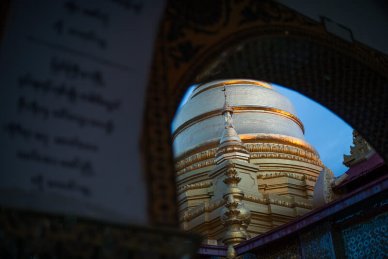 Gilded stupa of Sutaungpyei Pagoda framed through ornate arch, catching midday light at summit of Mandalay Hill
