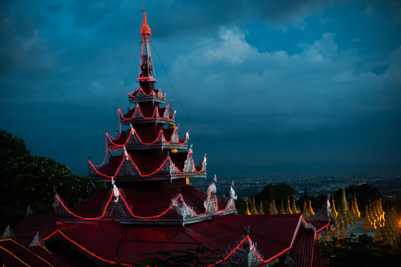 Red-lit pyatthat tower of Sutaungpyei Pagoda against stormy dusk sky, row of golden stupas along ridge to right