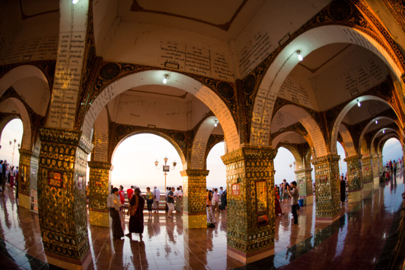 Glass-mosaic columns carrying arched portico at Sutaungpyei Pagoda, Burmese-script inscriptions on white vaults, crowd gathering for sunset