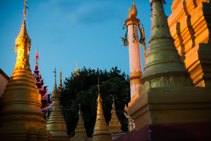Multiple gilded stupas, red pavilion roof, and hintha-topped column crowding Sutaungpyei Pagoda summit terrace at twilight
