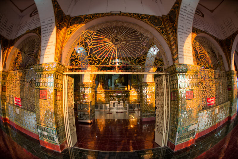 Gilded wrought-iron screen and glass-mosaic door jambs framing shrine entrance at Sutaungpyei Pagoda, seated Buddha visible in lamplight beyond
