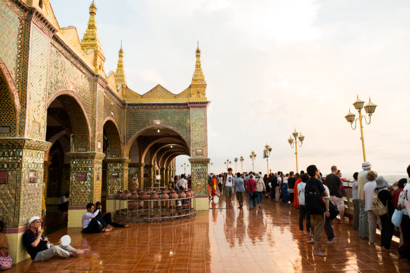 Barefoot crowds on Sutaungpyei Pagoda sunset terrace, glass-mosaic shrine hall and gilded spirelets rising to left, water jars in foreground