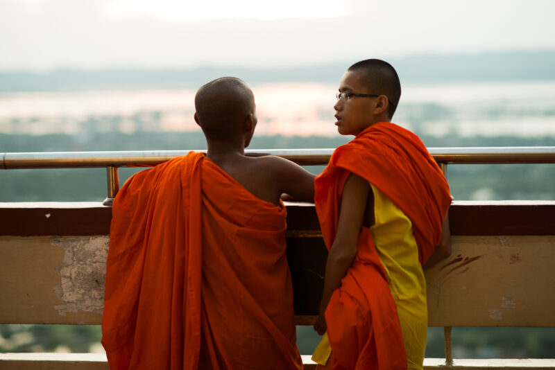 Two novice monks in orange robes leaning against terrace railing at Sutaungpyei Pagoda, Ayeyarwady River shimmering in haze below