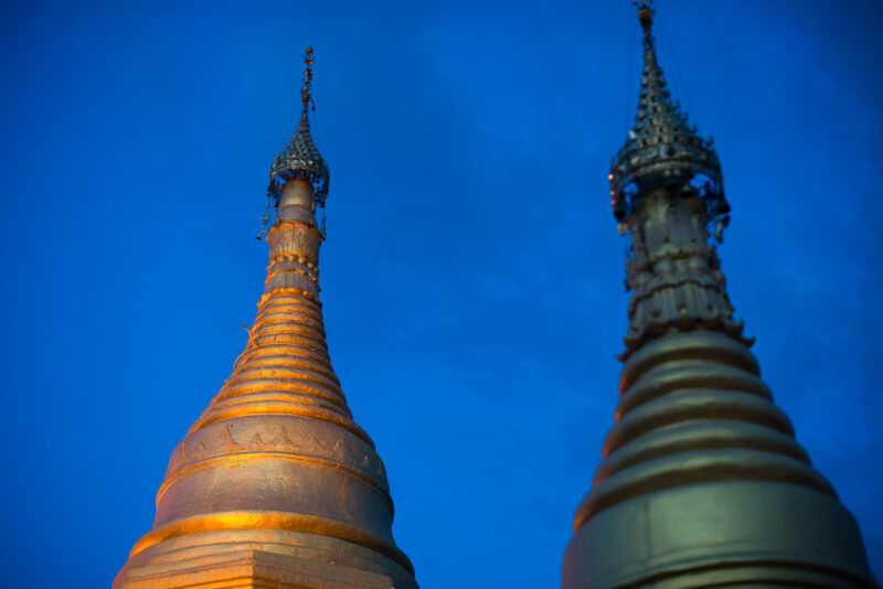 Two illuminated stupas at Sutaungpyei Pagoda against deep blue twilight, tiered bases and ornate metal hti finials outlined by light