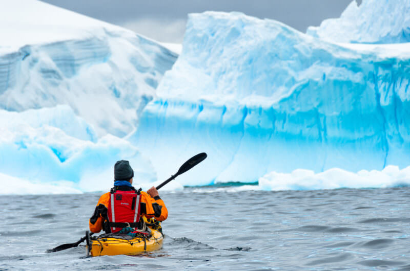 Sea kayaker in drysuit paddling toward massive blue iceberg at Melchior Islands, Antarctic Peninsula